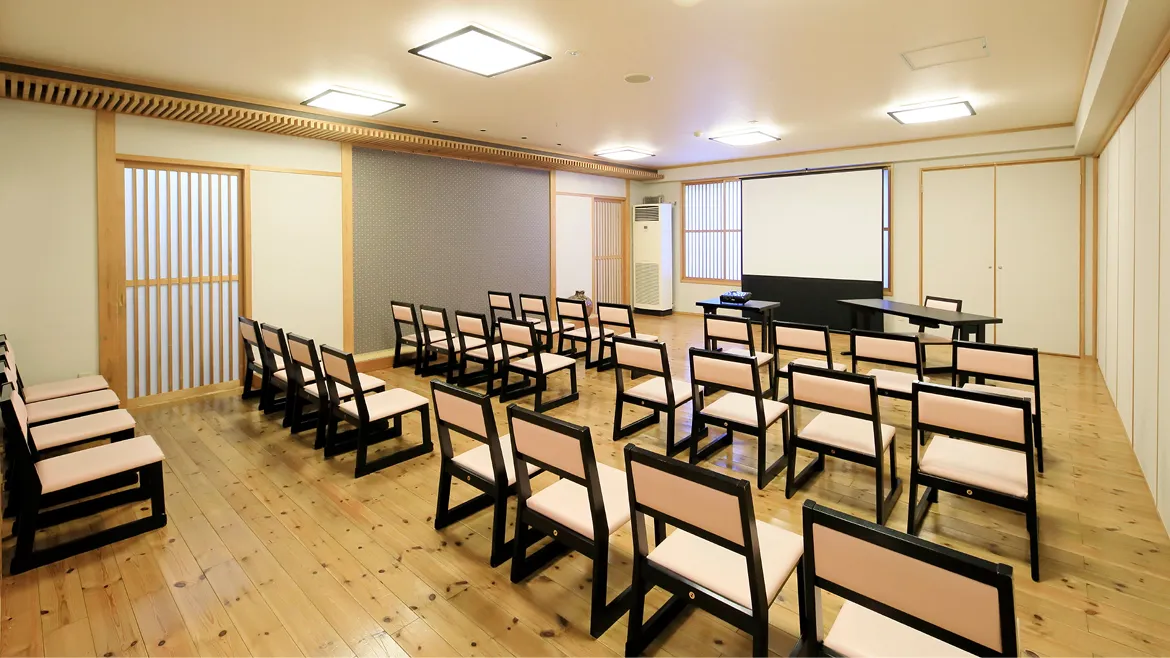 和室の大広間。正面にスクリーンと演台、多数の椅子が整然と並べられている。