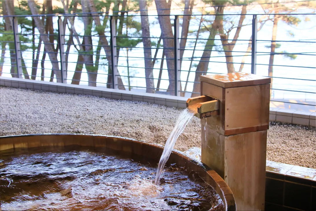 湯気が立ち上る露天風呂。中央には湯口があり、湯が勢いよく浴槽に注ぎ込まれている。