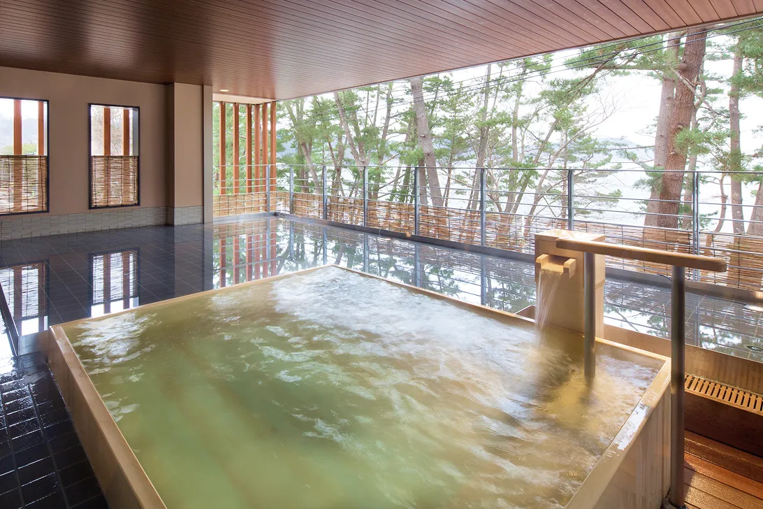 木造りの露天風呂。湯気が立ち上る浴槽の向こうには、緑豊かな木々が広がる。