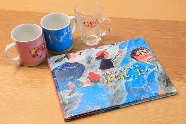 テーブルの上に、2つのマグカップと1つのグラス、絵本『つなみてんでんこ はしれ、上へ！』が置かれている。