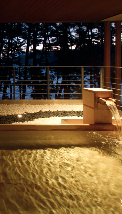 石造りの浴槽から湯が溢れ出て、湯煙が立ち上る露天風呂。背景には木々が生い茂り、夕焼け空が広がっています。