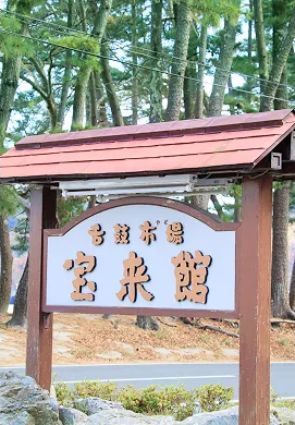 緑豊かな松林の中に立つ、木製の立て看板。看板には『名湯温泉 宝来館』と書かれている