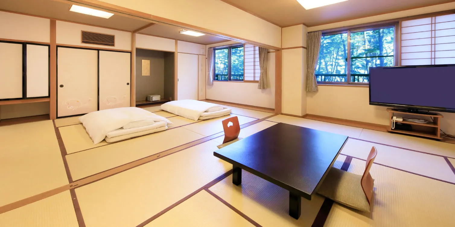 畳敷きの客室で、布団2つが用意されています。黒いローテーブルとテレビがあり、窓の外には緑豊かな景色が広がっています。