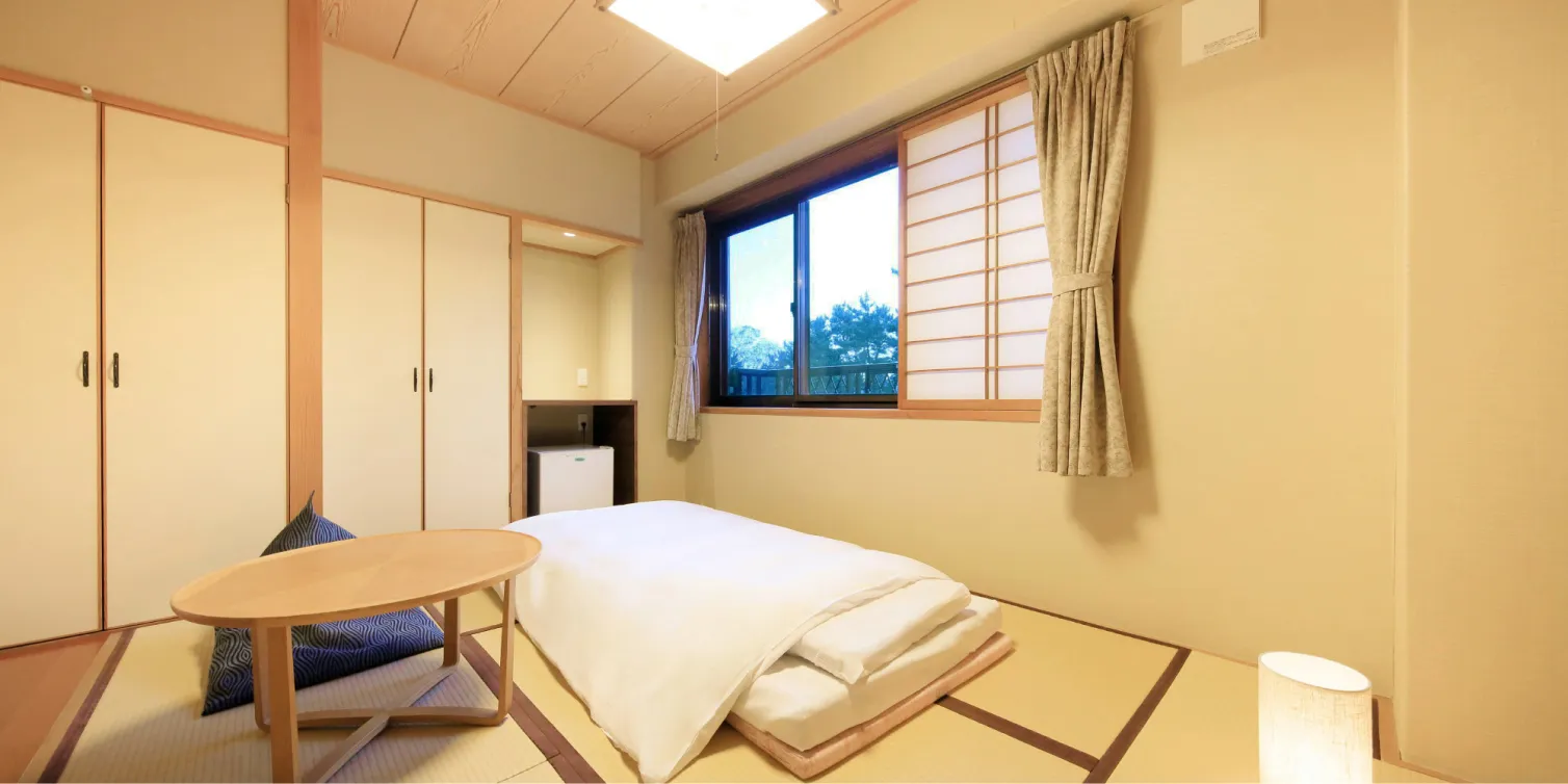 窓には障子とカーテンが掛けられている。部屋の中央にはベッドが置かれ、その横に小さなテーブルと照明器具が配置されている。